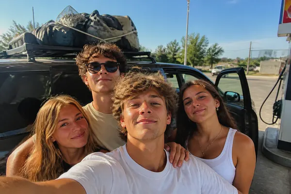 four friends squeezed into a selfie beside a loaded car at a petrol stop